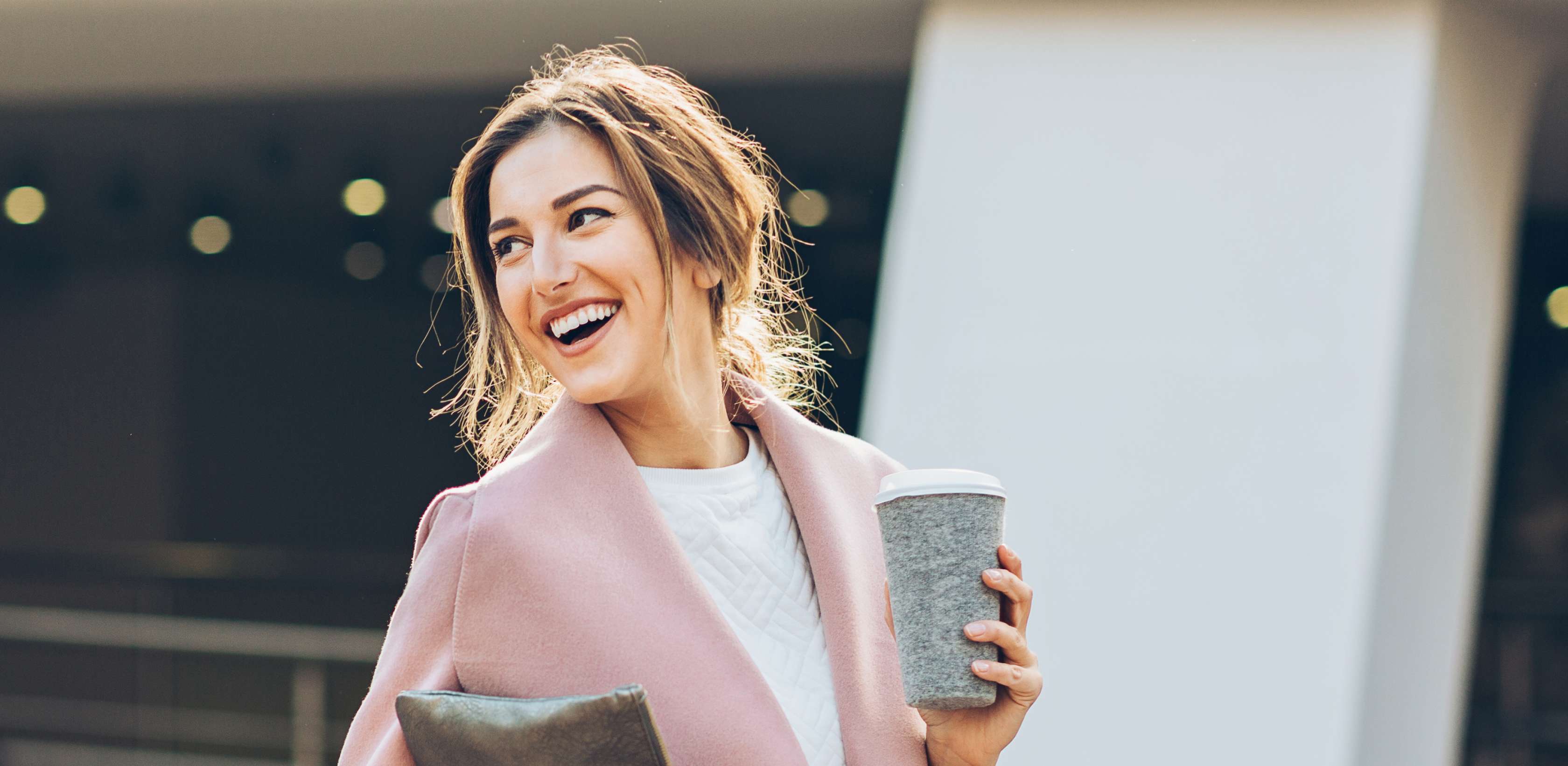 Junge lachende Frau in einem rosafarbenen Mantel hält einen Coffee-to-go-Becher